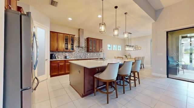 kitchen with stainless steel fridge, an island with sink, tasteful backsplash, pendant lighting, and wall chimney exhaust hood