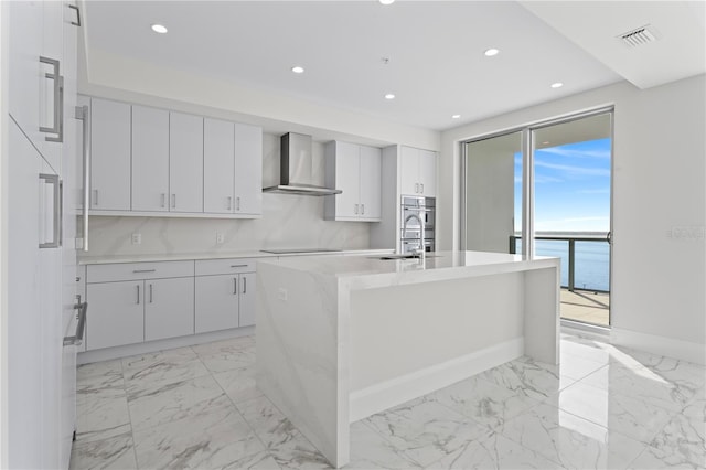 kitchen featuring white cabinetry, wall chimney exhaust hood, tasteful backsplash, a center island with sink, and a water view