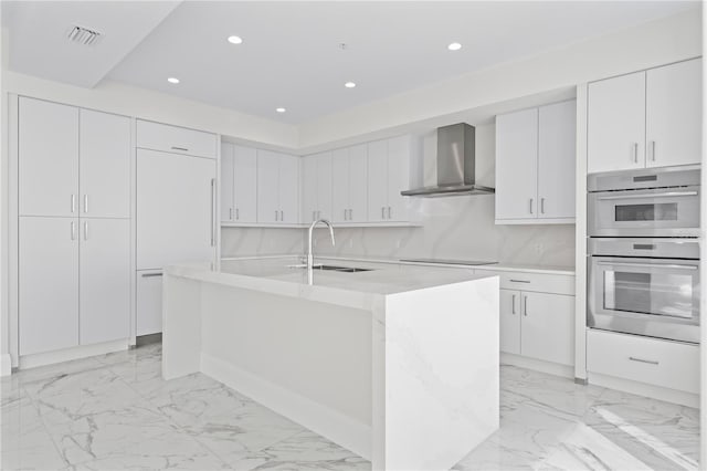 kitchen with sink, wall chimney exhaust hood, stainless steel double oven, a center island with sink, and white cabinets