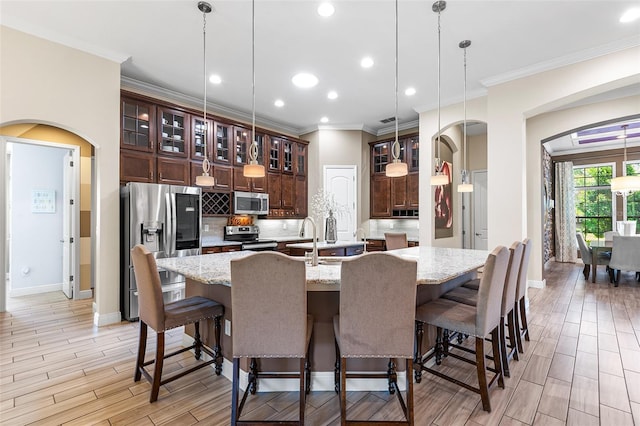 kitchen with light stone countertops, hanging light fixtures, an island with sink, appliances with stainless steel finishes, and ornamental molding