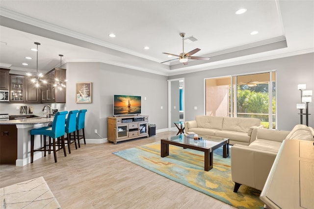 living room with ceiling fan, sink, crown molding, a tray ceiling, and light wood-type flooring