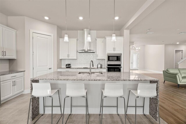 kitchen featuring stainless steel appliances, white cabinetry, hanging light fixtures, a kitchen island with sink, and wall chimney range hood