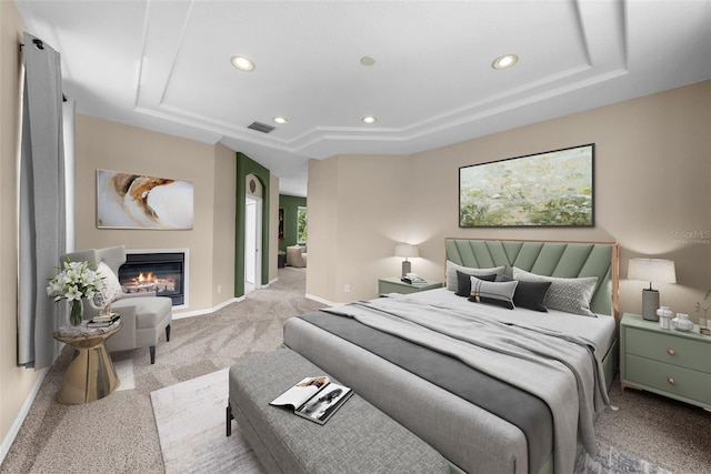 bedroom featuring recessed lighting, a raised ceiling, a glass covered fireplace, light carpet, and baseboards