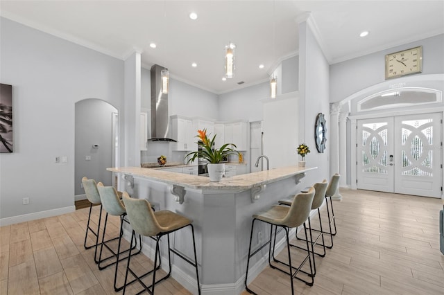 kitchen with wall chimney range hood, light stone counters, ornamental molding, white cabinets, and a kitchen bar