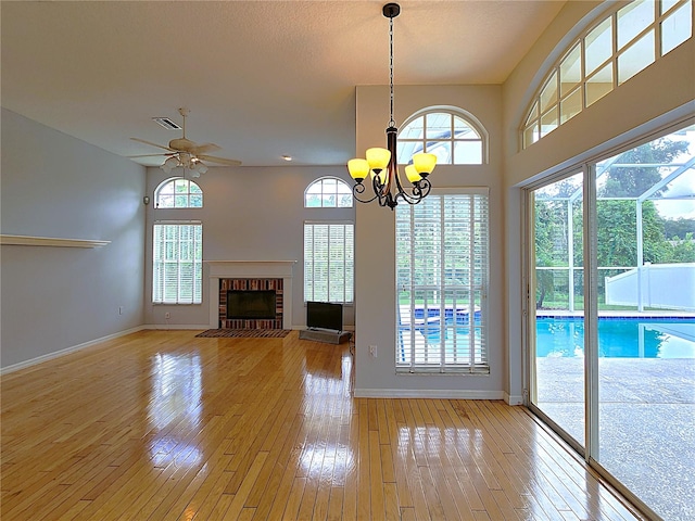 unfurnished living room featuring ceiling fan with notable chandelier, a brick fireplace, light wood-style floors, and baseboards