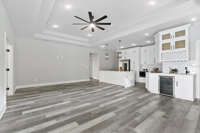 unfurnished living room with indoor wet bar, wine cooler, ceiling fan, a tray ceiling, and light wood-type flooring