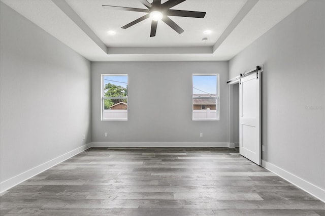 unfurnished bedroom with wood-type flooring, a barn door, ceiling fan, and a tray ceiling