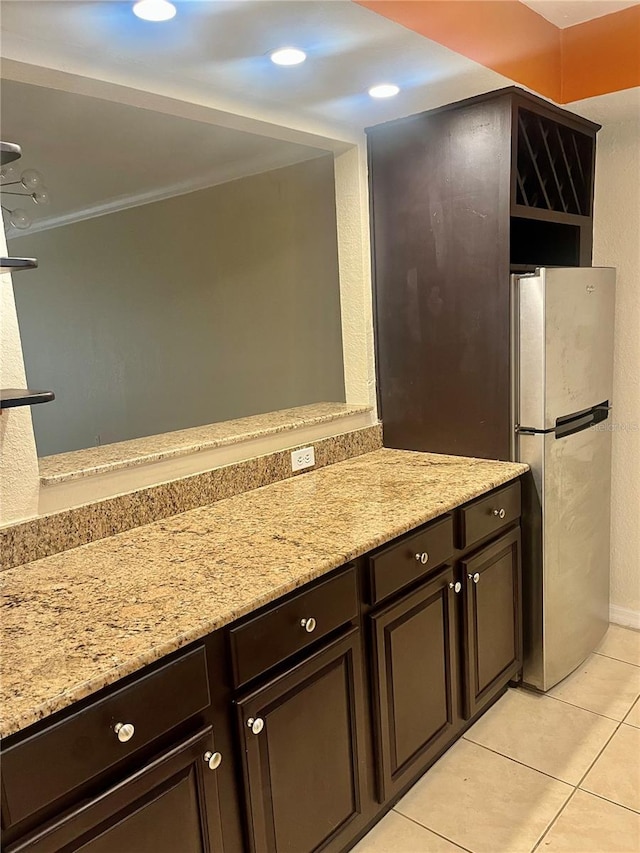 kitchen with light tile patterned flooring, stainless steel fridge, ornamental molding, dark brown cabinetry, and light stone counters