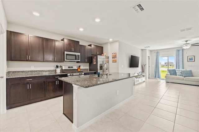 kitchen with appliances with stainless steel finishes, dark stone counters, a center island with sink, light tile patterned floors, and ceiling fan