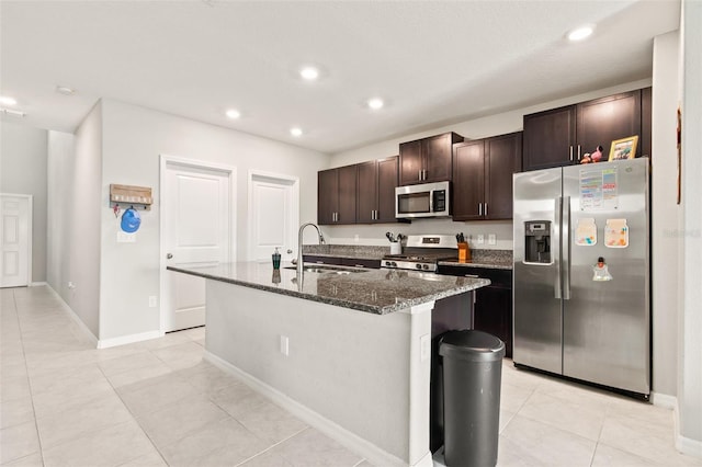 kitchen with stainless steel appliances, sink, dark stone countertops, light tile patterned floors, and a kitchen island with sink