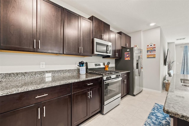 kitchen with appliances with stainless steel finishes, light tile patterned flooring, light stone countertops, and dark brown cabinetry