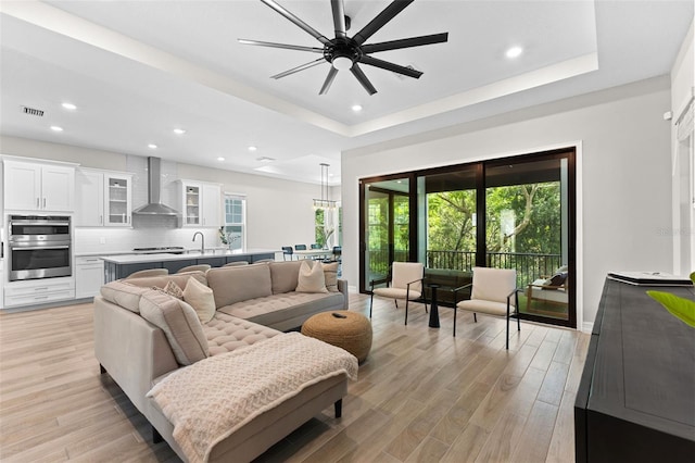 living room featuring ceiling fan, sink, light wood-type flooring, and a tray ceiling