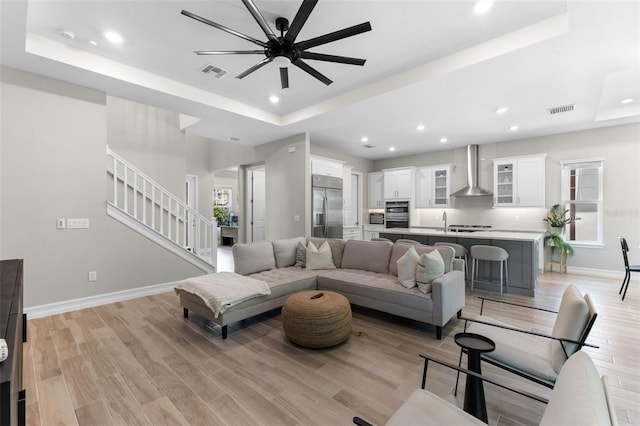 living room with ceiling fan, a tray ceiling, sink, and light hardwood / wood-style flooring