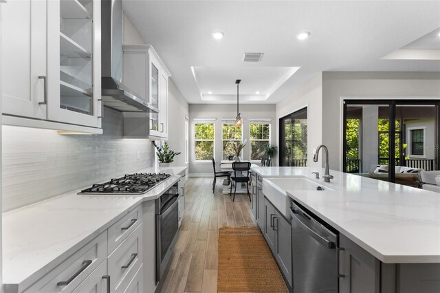 kitchen with decorative light fixtures, a raised ceiling, an island with sink, stainless steel appliances, and wall chimney exhaust hood