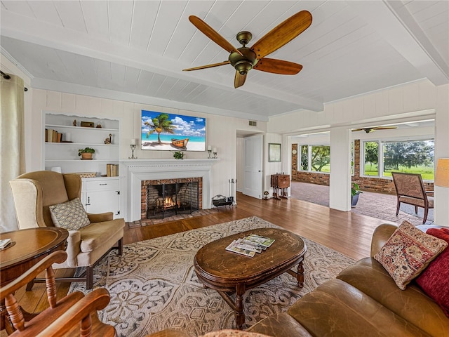 living room featuring ceiling fan, a fireplace, wood-type flooring, beam ceiling, and built in shelves