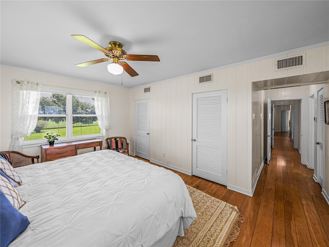 bedroom featuring dark wood-type flooring, two closets, wooden walls, ceiling fan, and crown molding