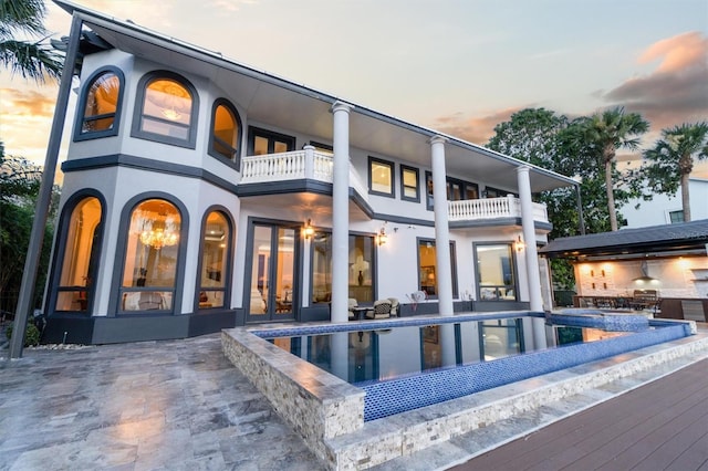 back of house at dusk with an infinity pool, stucco siding, a patio area, a balcony, and exterior kitchen