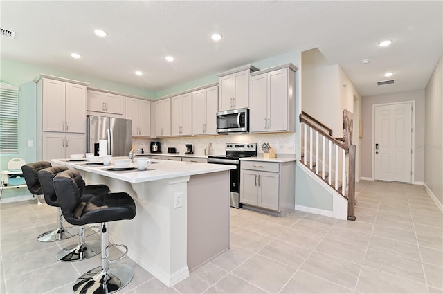 kitchen featuring a center island with sink, stainless steel appliances, light tile patterned flooring, a breakfast bar, and backsplash