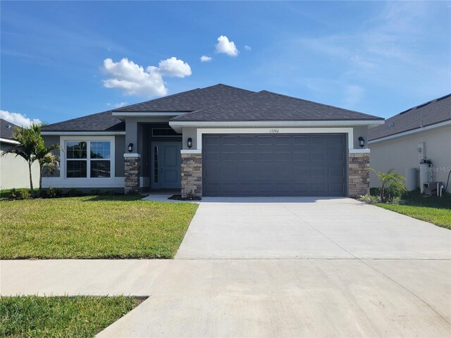 prairie-style home featuring stucco siding, a front yard, stone siding, an attached garage, and driveway