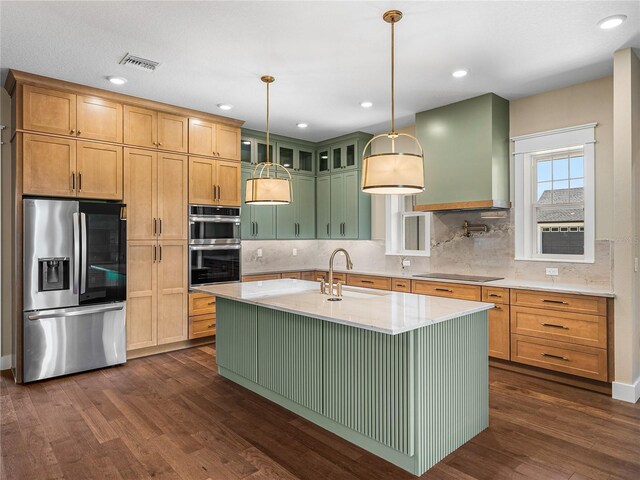 kitchen featuring wall chimney range hood, dark hardwood / wood-style flooring, decorative backsplash, an island with sink, and appliances with stainless steel finishes