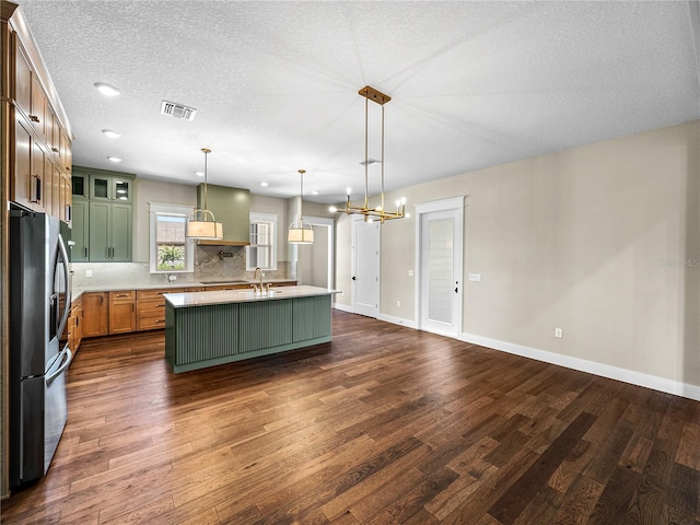 kitchen with hanging light fixtures, stainless steel fridge with ice dispenser, wall chimney exhaust hood, dark hardwood / wood-style floors, and a kitchen island with sink