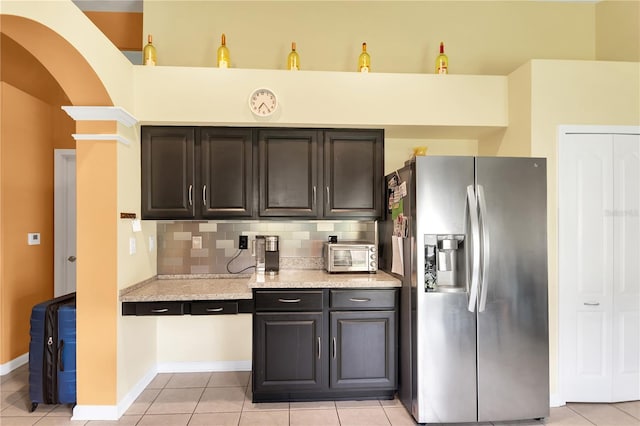 kitchen with dark brown cabinets, stainless steel fridge, backsplash, light tile patterned floors, and light stone counters