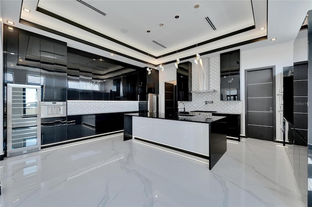 kitchen with wall chimney exhaust hood, sink, backsplash, a tray ceiling, and a center island with sink