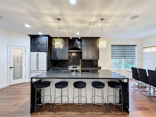 kitchen with a breakfast bar area, decorative light fixtures, a center island with sink, and wall chimney range hood