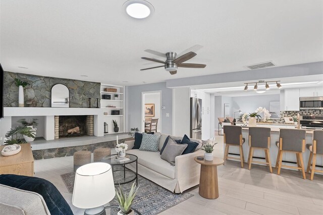 living room with built in features, a stone fireplace, ceiling fan, and track lighting