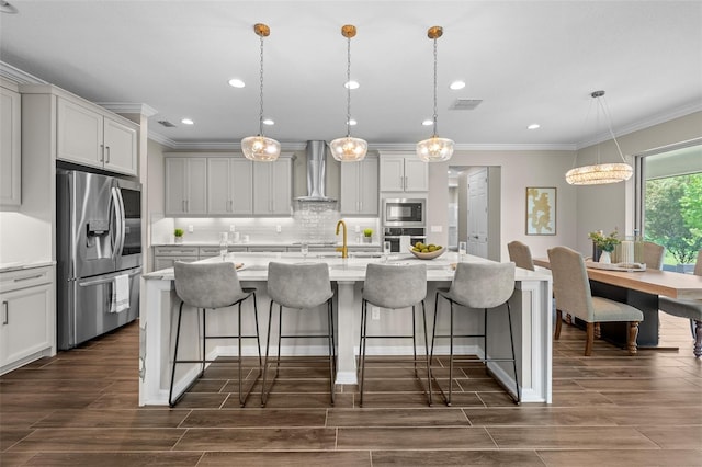 kitchen featuring decorative light fixtures, a kitchen island with sink, appliances with stainless steel finishes, and wall chimney range hood