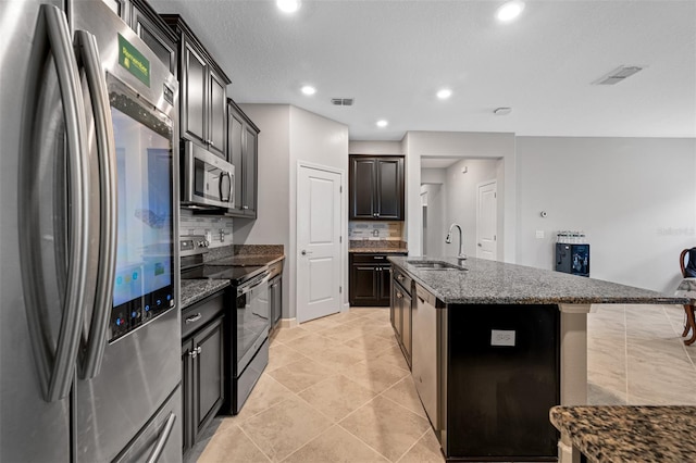 kitchen featuring stainless steel appliances, a center island with sink, dark stone countertops, backsplash, and light tile patterned flooring