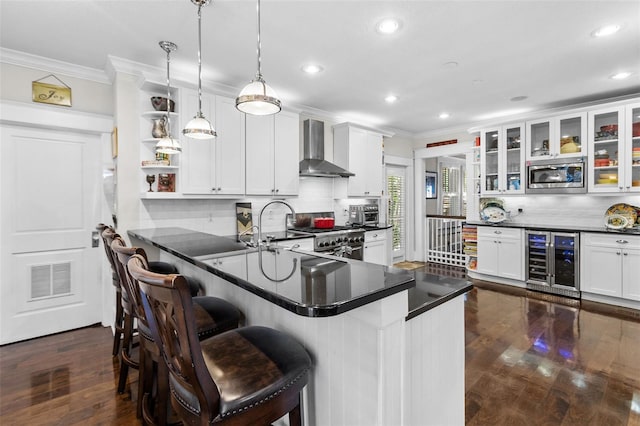 kitchen featuring wall chimney exhaust hood, a kitchen bar, appliances with stainless steel finishes, pendant lighting, and beverage cooler