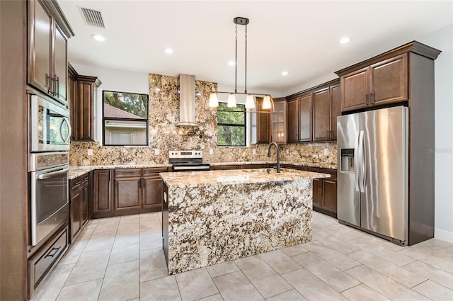 kitchen with hanging light fixtures, a kitchen island with sink, sink, stainless steel appliances, and wall chimney exhaust hood