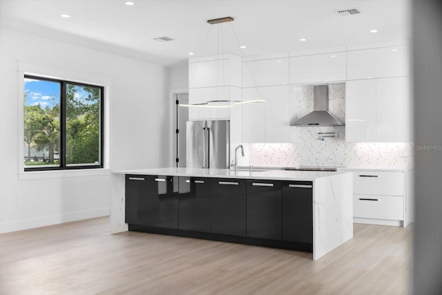 kitchen featuring white cabinets, decorative light fixtures, wall chimney range hood, a center island with sink, and high end refrigerator