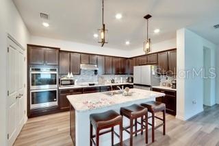 kitchen featuring dark brown cabinets, white refrigerator, pendant lighting, stainless steel double oven, and a kitchen island with sink