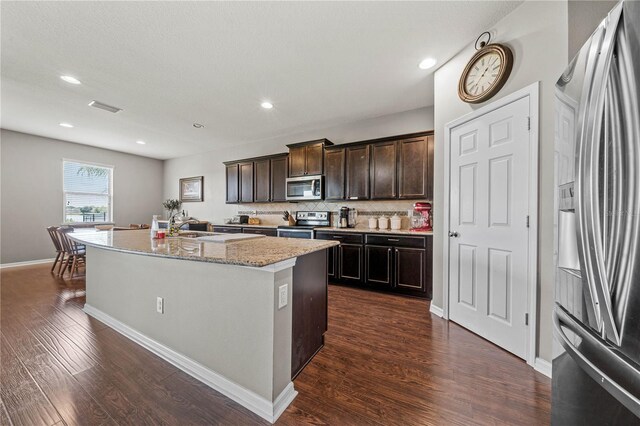 kitchen featuring dark brown cabinets, stainless steel appliances, an island with sink, light stone counters, and dark wood-type flooring