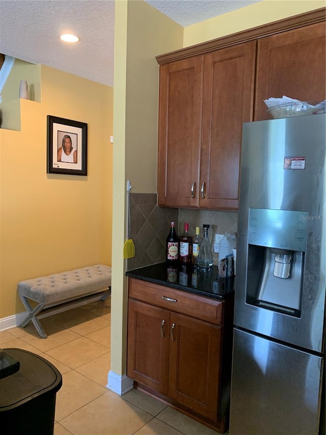 kitchen featuring a textured ceiling, light tile patterned flooring, backsplash, dark stone countertops, and stainless steel fridge