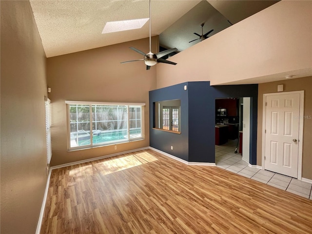 unfurnished living room featuring a skylight, ceiling fan, light wood-type flooring, high vaulted ceiling, and a textured ceiling