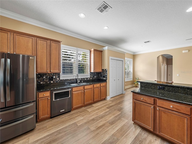 kitchen with stainless steel appliances, dark stone counters, ornamental molding, sink, and light wood-type flooring