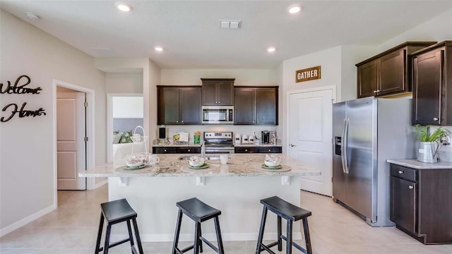 kitchen with appliances with stainless steel finishes, a center island with sink, dark brown cabinets, light stone countertops, and light tile patterned flooring