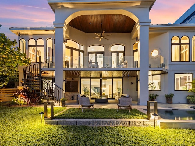 back house at dusk with a lawn, ceiling fan, outdoor lounge area, a patio area, and a balcony