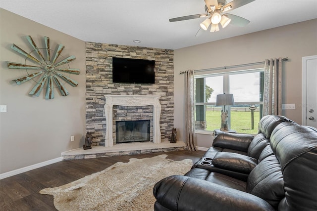 living area featuring a ceiling fan, a stone fireplace, baseboards, and wood finished floors