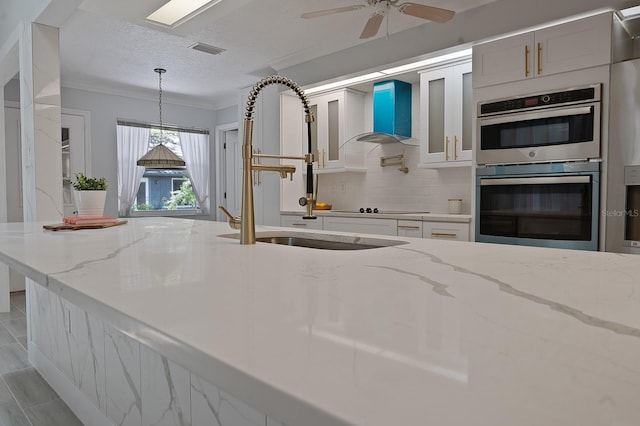 kitchen featuring ceiling fan, double oven, light stone countertops, wall chimney exhaust hood, and decorative light fixtures