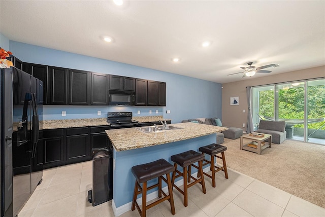 kitchen featuring black appliances, an island with sink, light stone counters, light colored carpet, and a breakfast bar area