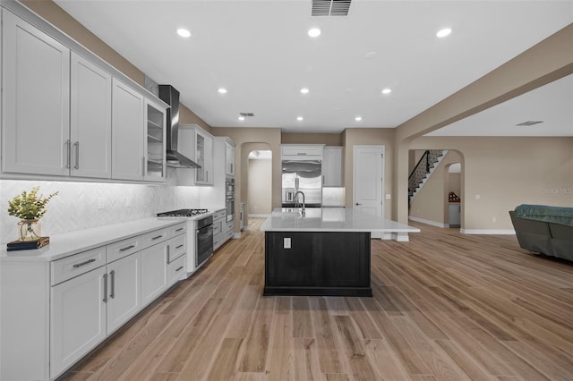 kitchen with a kitchen island with sink, light wood-type flooring, white cabinets, and wall chimney range hood