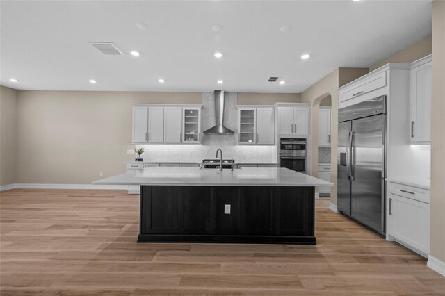 kitchen with an island with sink, wall chimney exhaust hood, light hardwood / wood-style flooring, and stainless steel appliances