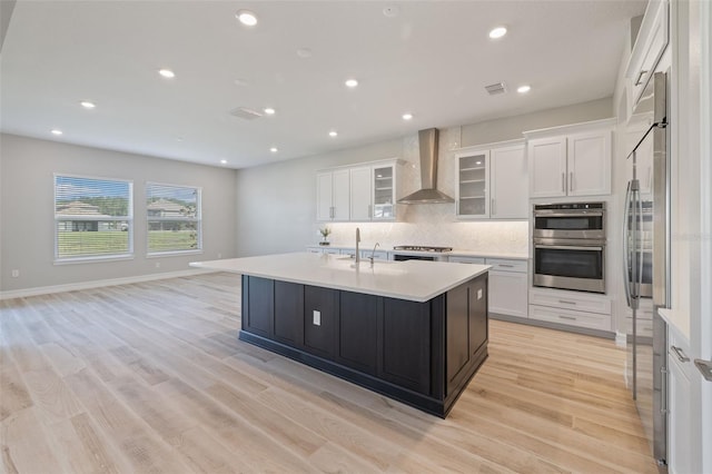 kitchen with an island with sink, stainless steel appliances, white cabinetry, and wall chimney range hood