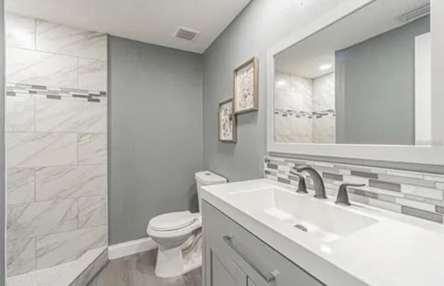 bathroom with toilet, visible vents, vanity, tiled shower, and decorative backsplash