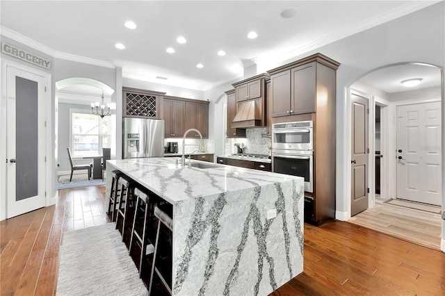 kitchen with sink, dark brown cabinets, stainless steel appliances, light stone countertops, and an island with sink