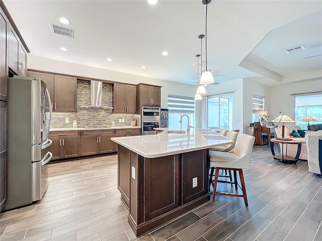 kitchen featuring pendant lighting, decorative backsplash, a kitchen island with sink, stainless steel appliances, and wall chimney exhaust hood